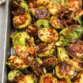 A baking tray filled with roasted Brussels sprouts, halved and crispy with a browned, seasoned coating. A checkered kitchen towel is partially visible in the background.