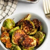 A white bowl filled with crispy, roasted Brussels sprouts, some with golden-brown, caramelized edges, sits on a light surface next to a gold fork and a checkered kitchen towel.