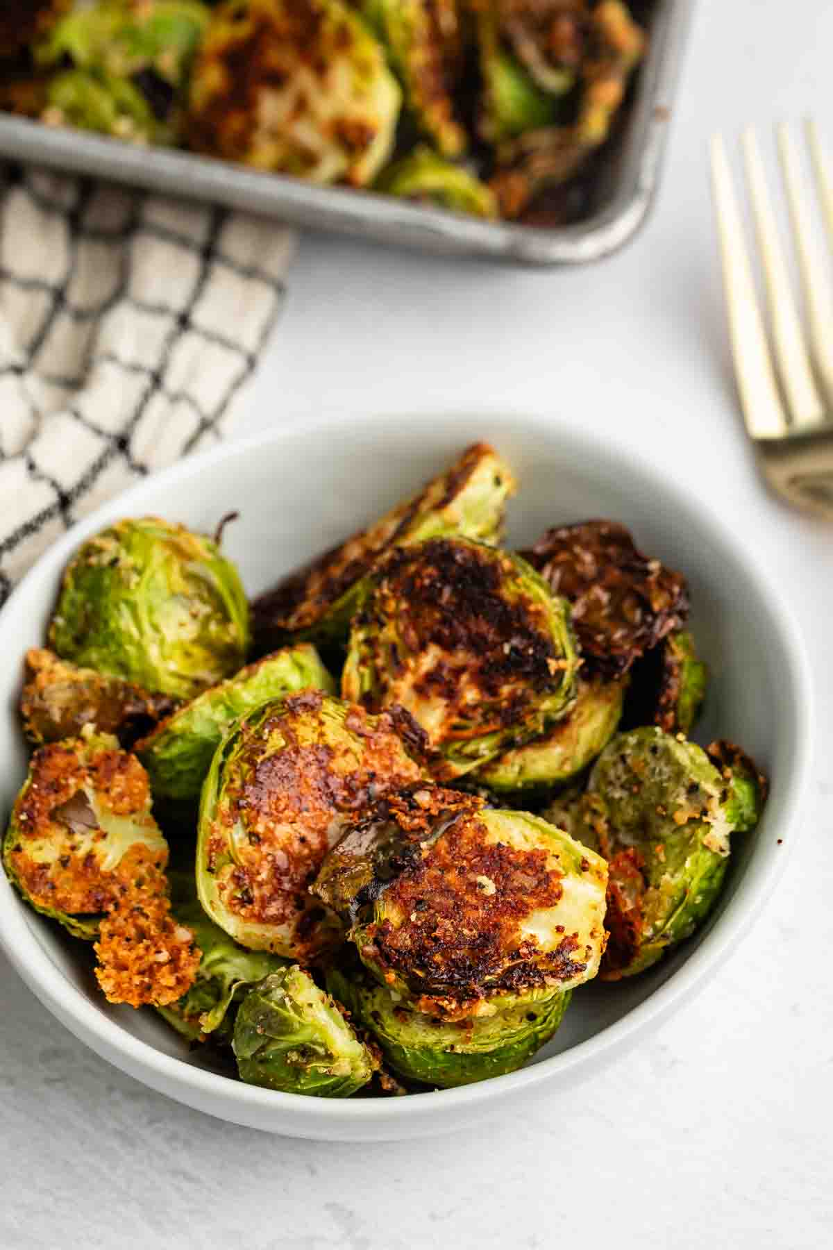 A white bowl filled with crispy, roasted Brussels sprouts, some with golden-brown, caramelized edges, sits on a light surface next to a gold fork and a checkered kitchen towel.