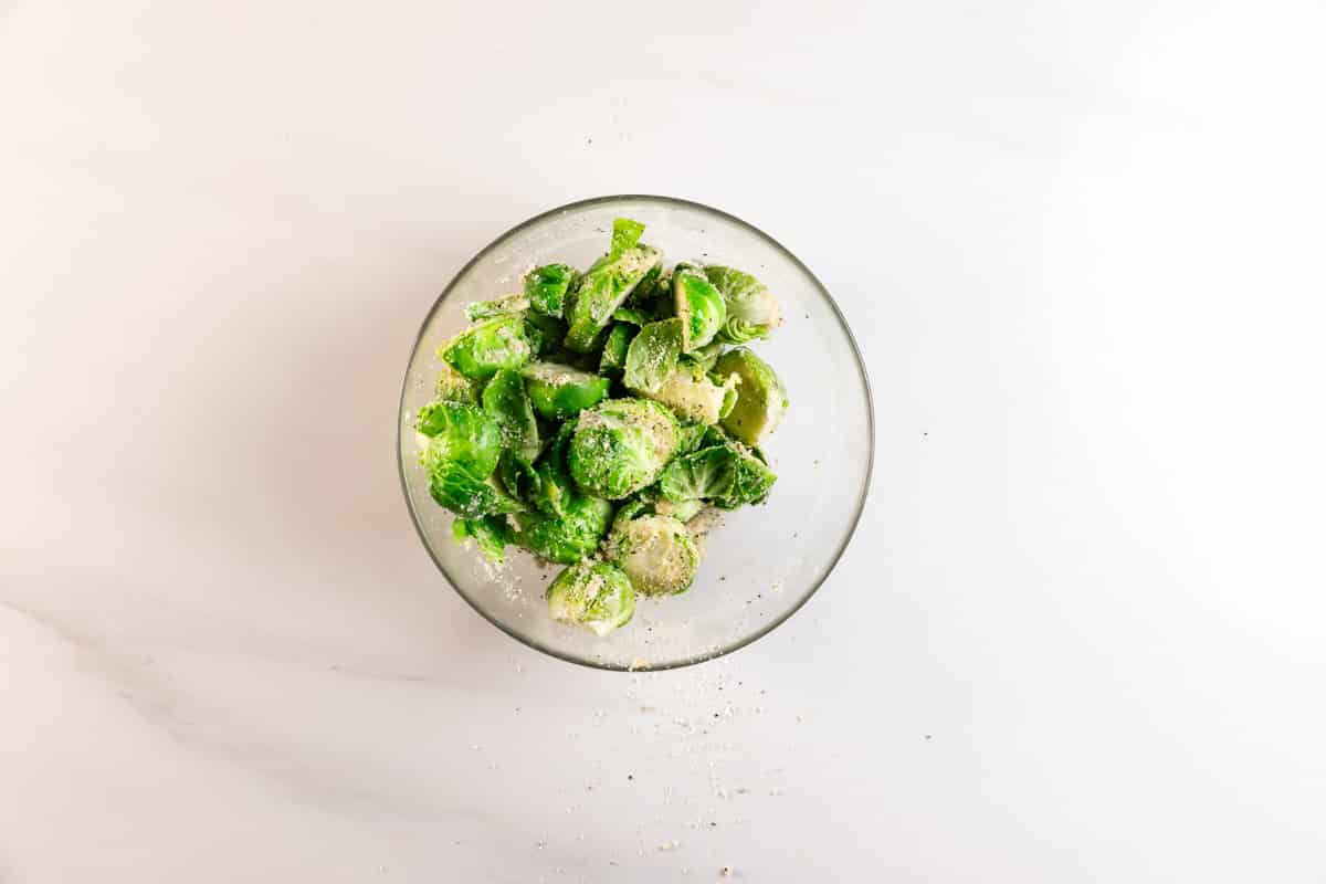 A clear glass bowl filled with raw Brussels sprouts sprinkled with grated cheese and pepper, placed on a white marble surface with some seasoning scattered around.