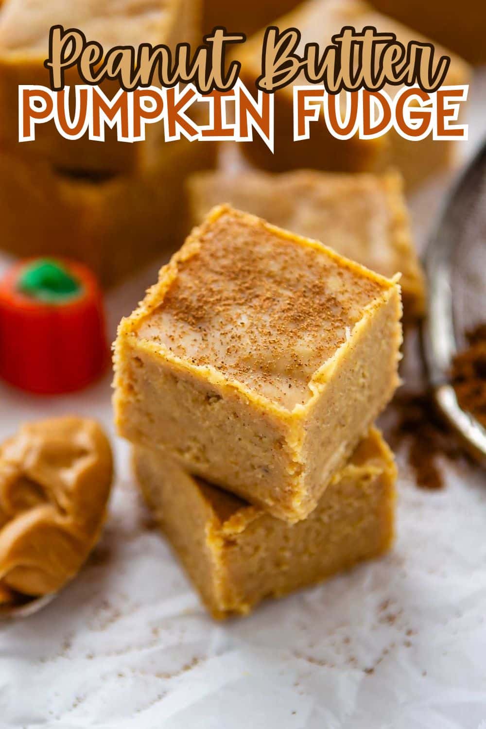 Two square pieces of peanut butter pumpkin fudge stacked on parchment paper, dusted with cinnamon, with a scoop of peanut butter and a small pumpkin candy nearby. Text above reads “Peanut Butter Pumpkin Fudge.”.