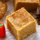 A close-up of a pumpkin spice-flavored dessert bar topped with a sprinkle of cinnamon, set on parchment paper with a small pumpkin-shaped candy nearby.