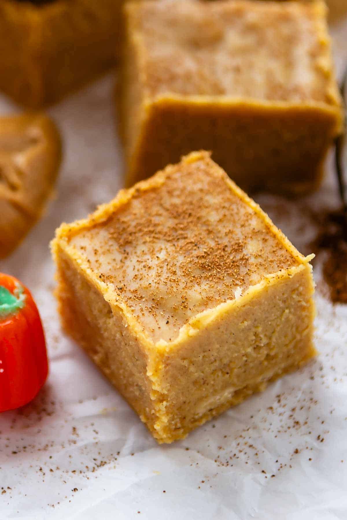 A close-up of a pumpkin spice-flavored dessert bar topped with a sprinkle of cinnamon, set on parchment paper with a small pumpkin-shaped candy nearby.