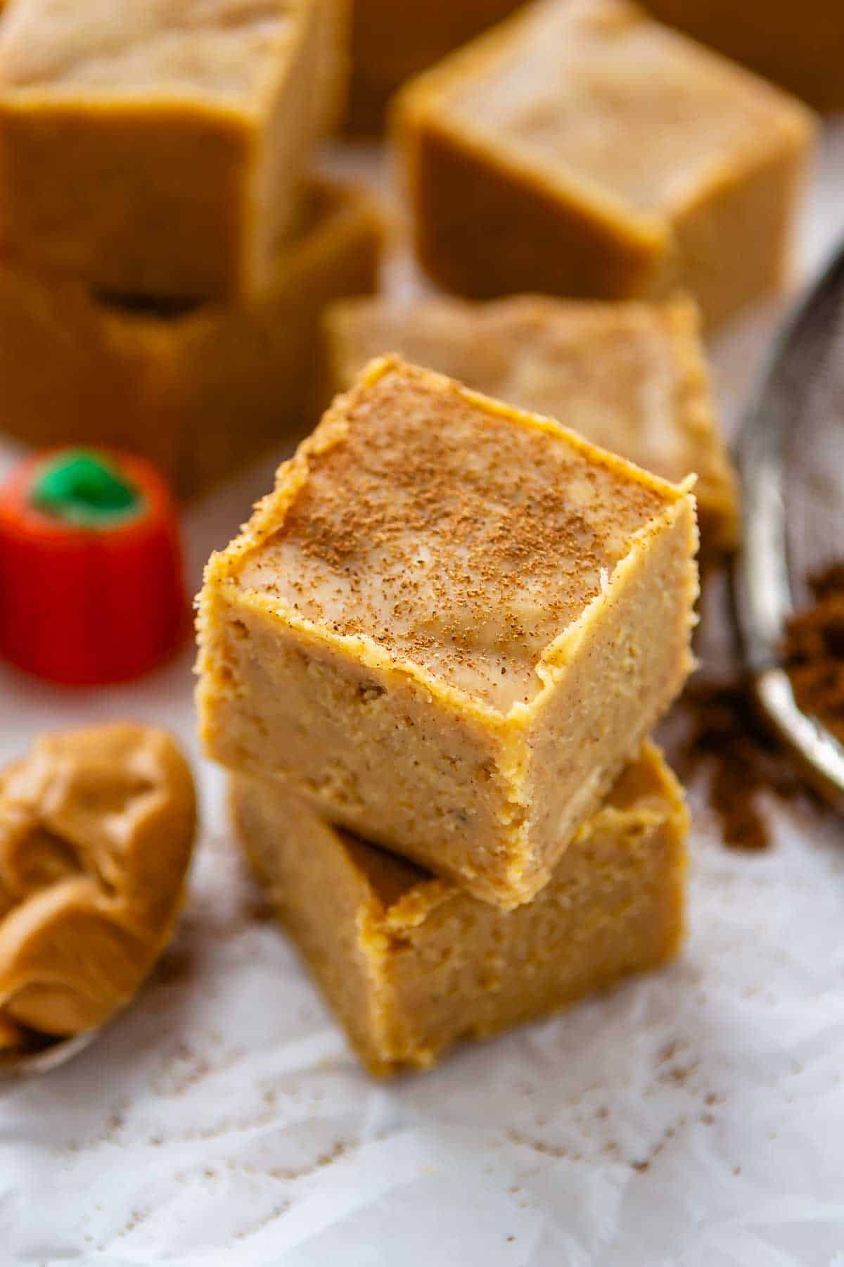 Two square pieces of blondie stacked on parchment paper, dusted with cinnamon, with more blondies, a candy pumpkin, and a spoonful of peanut butter in the background.