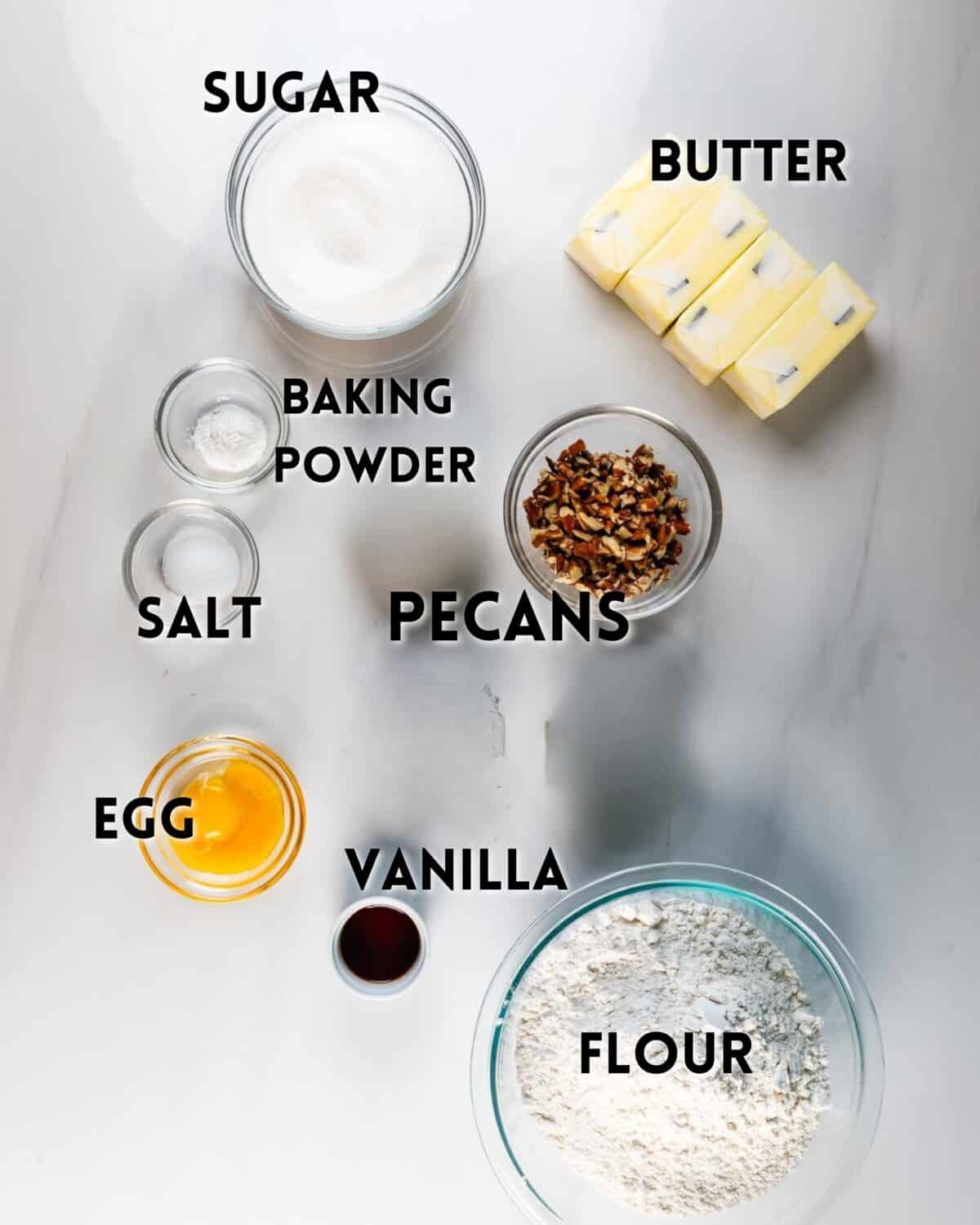 Overhead view of baking ingredients on a white surface, including labeled bowls of sugar, baking powder, salt, egg, vanilla, cranberries, pistachios, flour, and sliced butter.