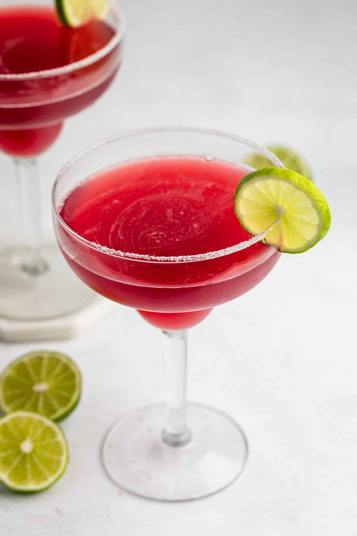 A margarita glass filled with a bright red cocktail, garnished with a lime wheel on the rim. Another similar glass and sliced limes are in the background on a white surface.