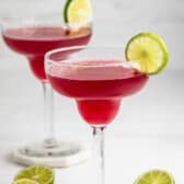Two margarita glasses filled with bright pink cocktail, each garnished with a slice of lime on the rim. There are halved limes on the white surface beside the glasses. The background is minimal and light.