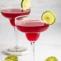 Two margarita glasses filled with bright pink cocktail, each garnished with a slice of lime on the rim. There are halved limes on the white surface beside the glasses. The background is minimal and light.