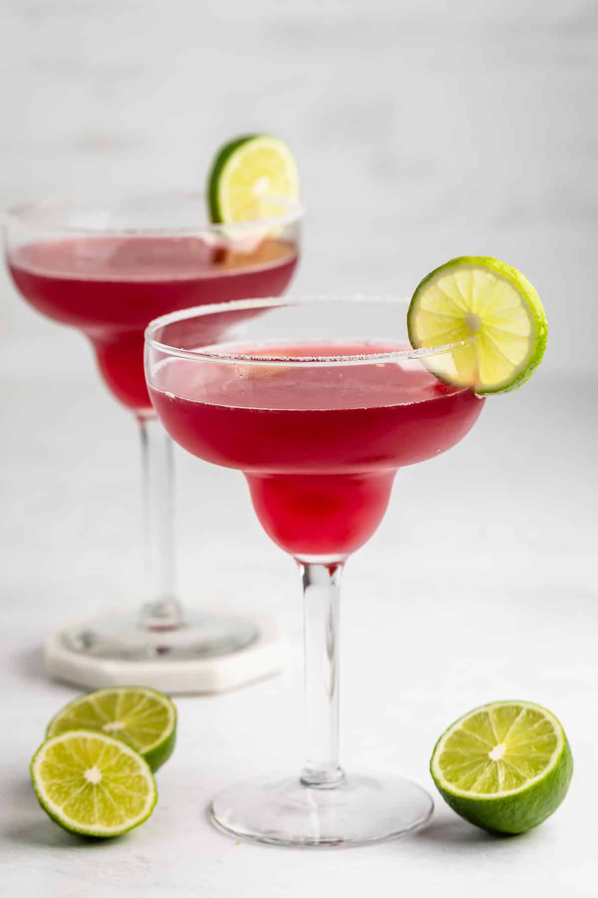 Two margarita glasses filled with bright pink cocktail, each garnished with a slice of lime on the rim. There are halved limes on the white surface beside the glasses. The background is minimal and light.