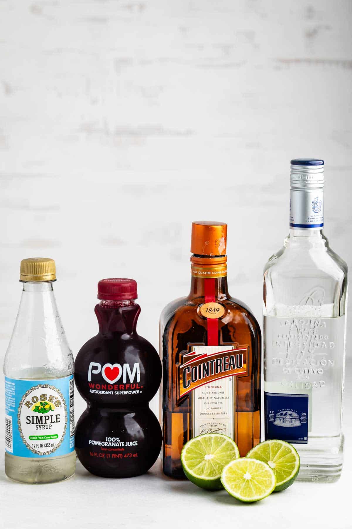 Four bottles (simple syrup, pomegranate juice, Cointreau, tequila) stand side by side on a white surface with three lime halves in front of them, set against a light background.