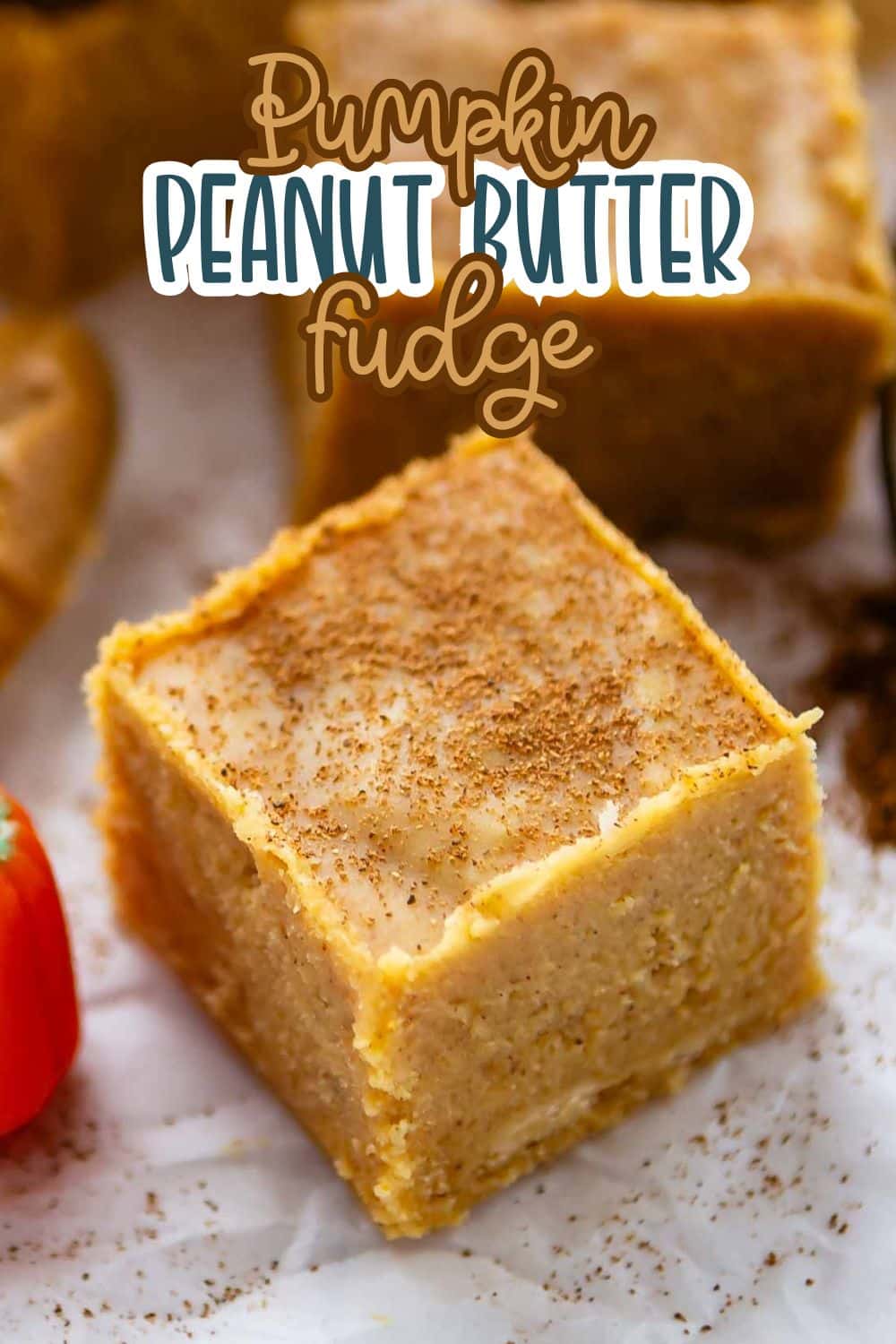 A close-up of a square piece of pumpkin peanut butter fudge on a white surface, lightly dusted with spices, with more fudge pieces in the background and text overlay that reads Pumpkin Peanut Butter Fudge.