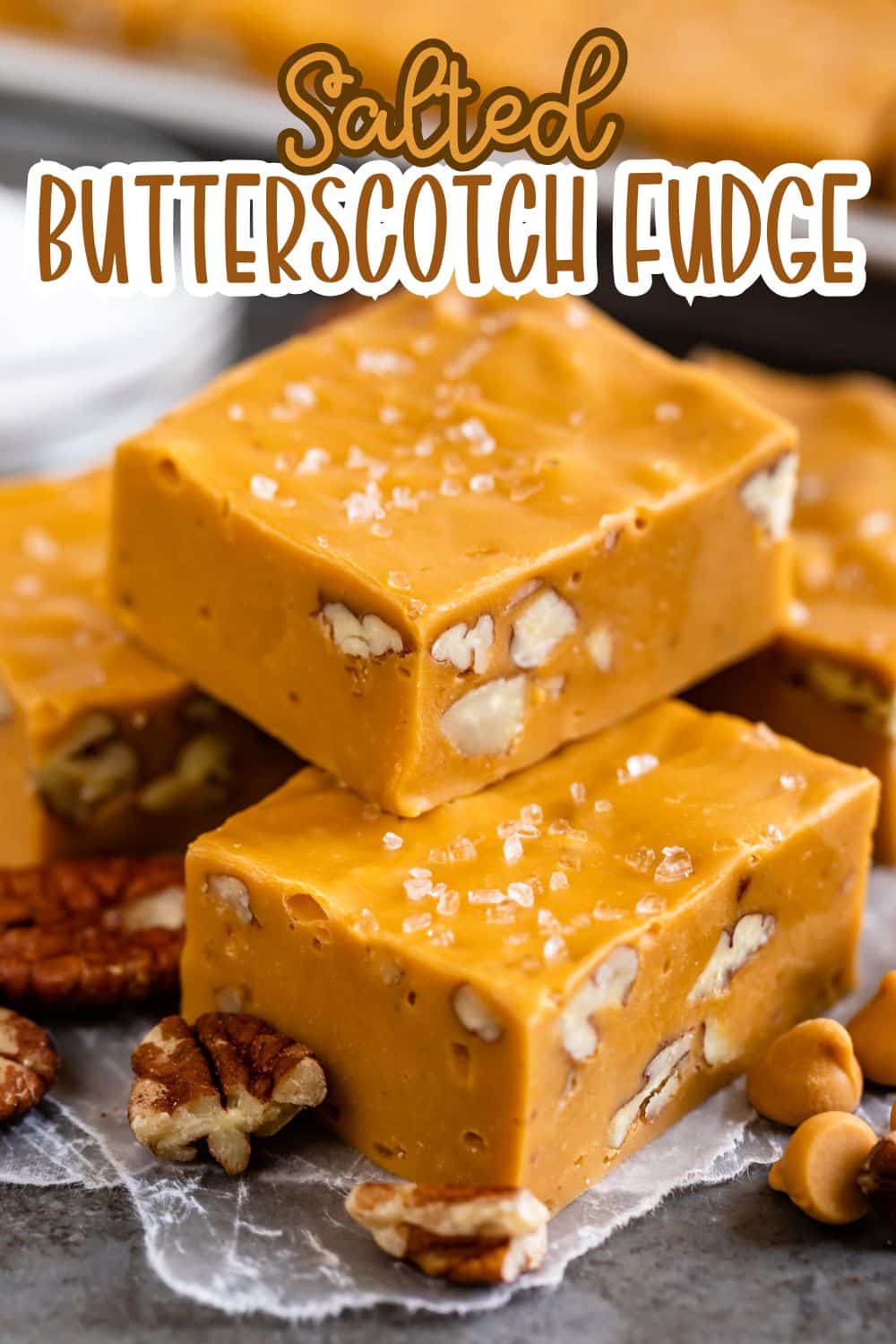 Three thick squares of salted butterscotch fudge with visible chopped pecans, topped with flaky sea salt, are stacked on a marble surface. Pecans and butterscotch chips are scattered around.