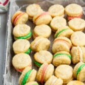 A tray of sandwich cookies with colorful cream fillings in green, pink, and white, arranged on parchment paper. Some cookies are stacked, and a striped towel is visible in the background.