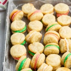 A tray of sandwich cookies with colorful cream fillings in green, pink, and white, arranged on parchment paper. Some cookies are stacked, and a striped towel is visible in the background.