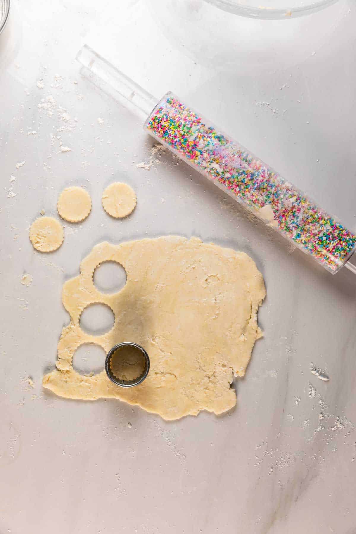 A sheet of rolled-out dough on a floured surface with three circles cut out, a round cookie cutter, and a rolling pin filled with colorful sprinkles nearby.