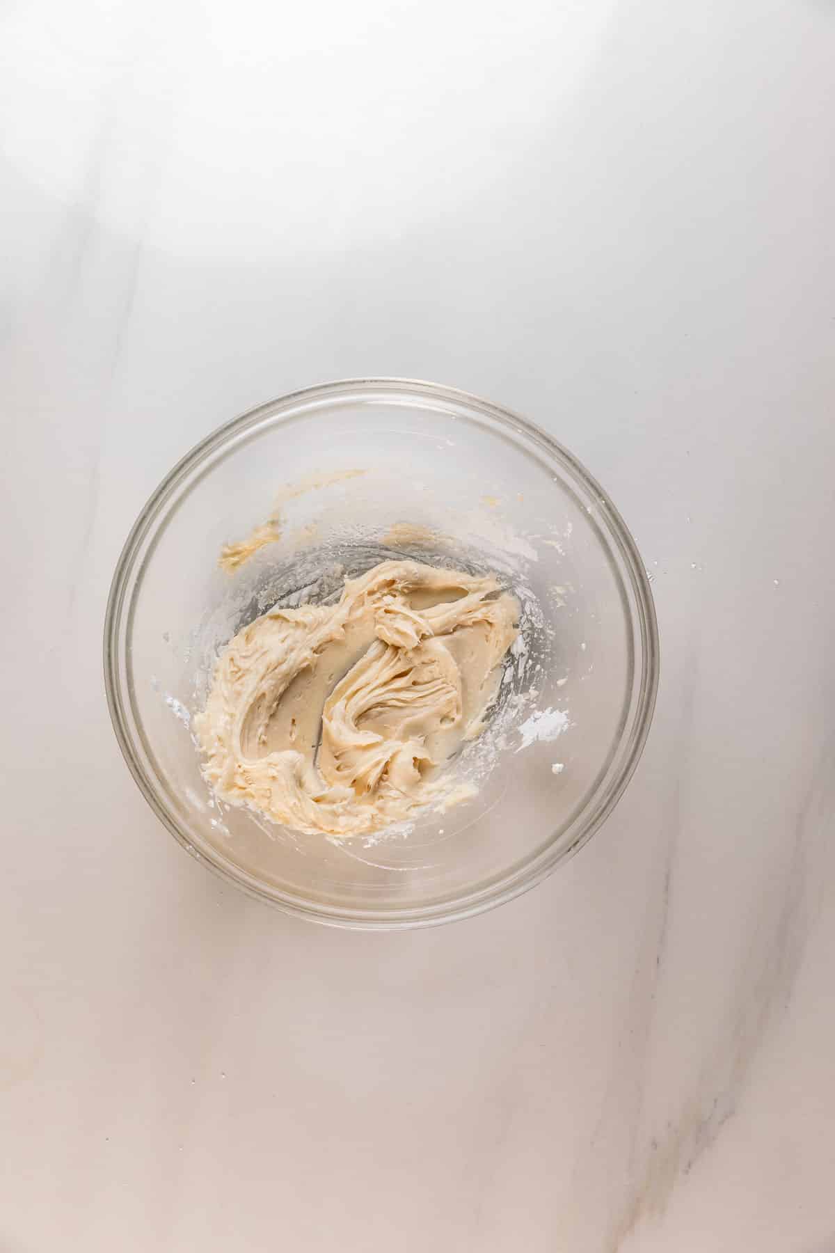 A clear glass bowl containing creamy, light tan dough or batter sits on a white marble surface. Some remnants of flour are visible around the dough inside the bowl.