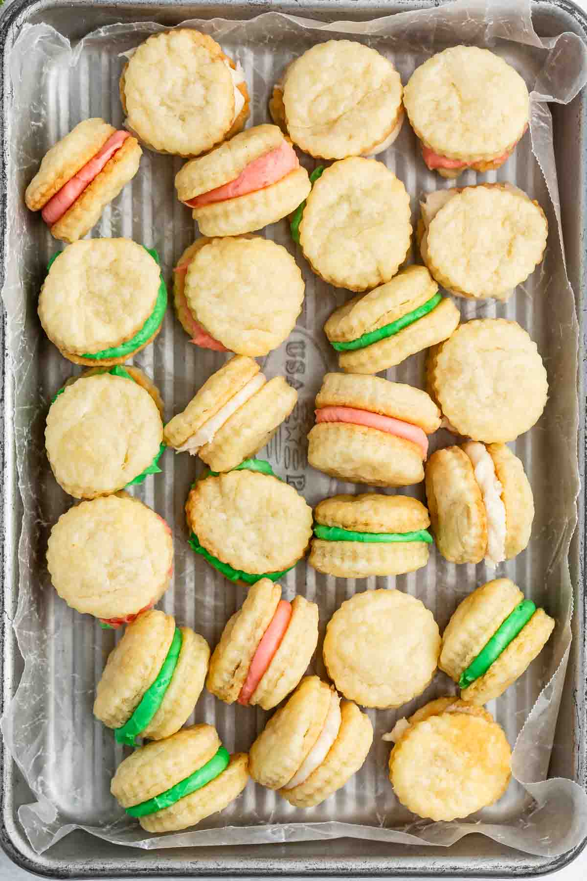 A tray lined with parchment holds sandwich cookies filled with green, pink, and white cream, arranged in a scattered pattern.
