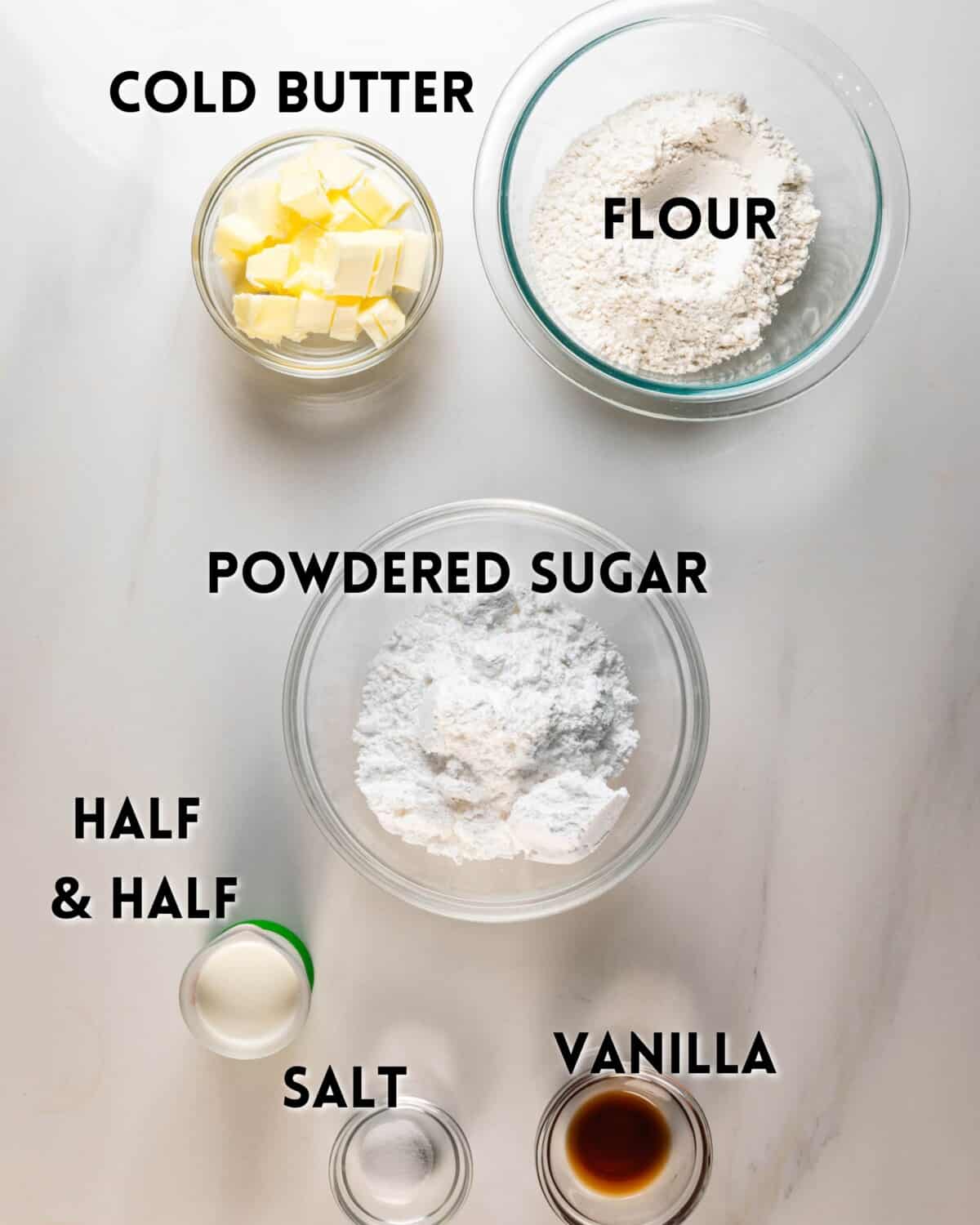 Five labeled bowls and jars of baking ingredients on a white surface: cubed cold butter, flour, powdered sugar, half & half, salt, and vanilla extract.