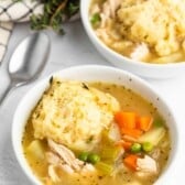 Two white bowls of chicken and vegetable soup with carrots, peas, and celery, each topped with a large dumpling. Fresh herbs and a spoon are nearby on a white surface.