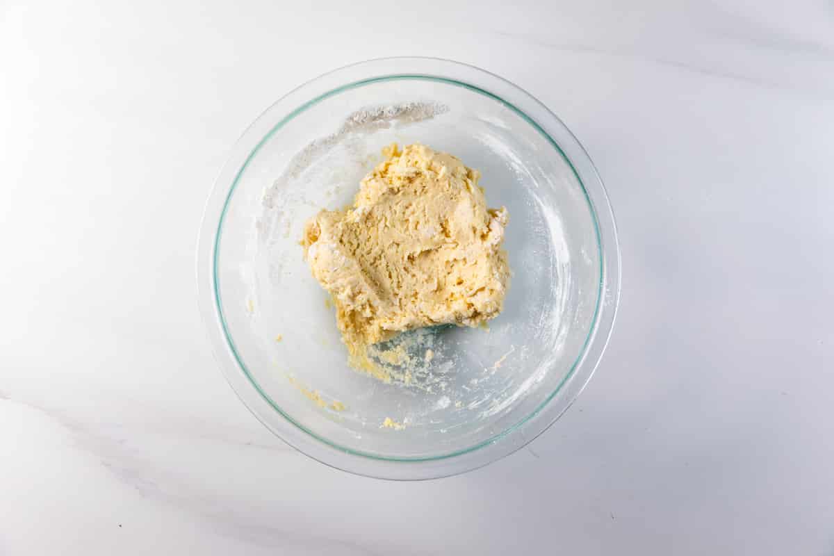 A glass bowl containing a ball of raw dough sits on a white marble surface. Some flour is scattered around the dough inside the bowl.