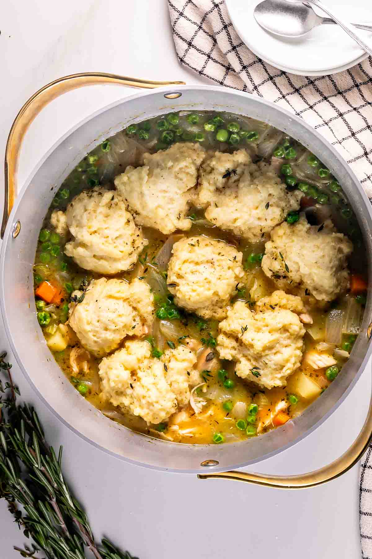 A pot of chicken and vegetable soup topped with fluffy dumplings, garnished with herbs. A striped towel, spoon, and plates are nearby, with sprigs of rosemary and thyme beside the pot.
