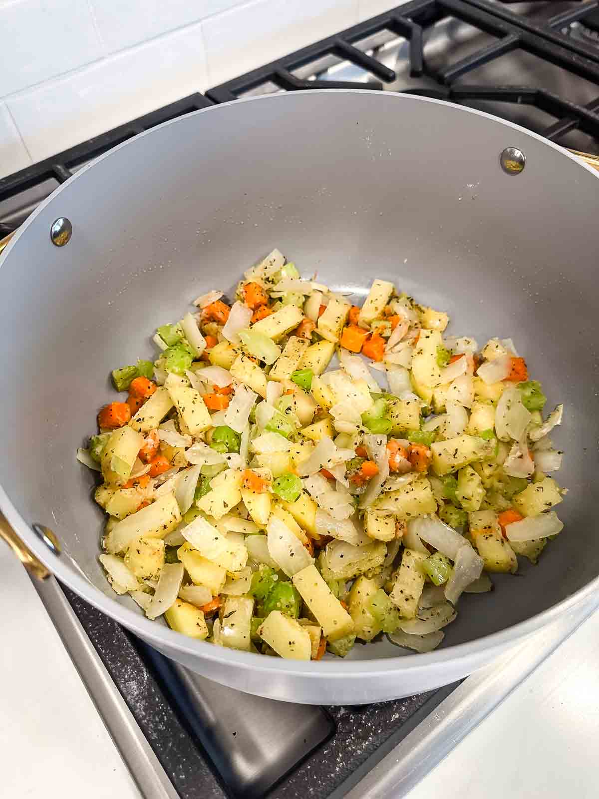 Chopped onions, carrots, celery, and potatoes sauté in a large gray pot on a stove, seasoned with herbs and black pepper.