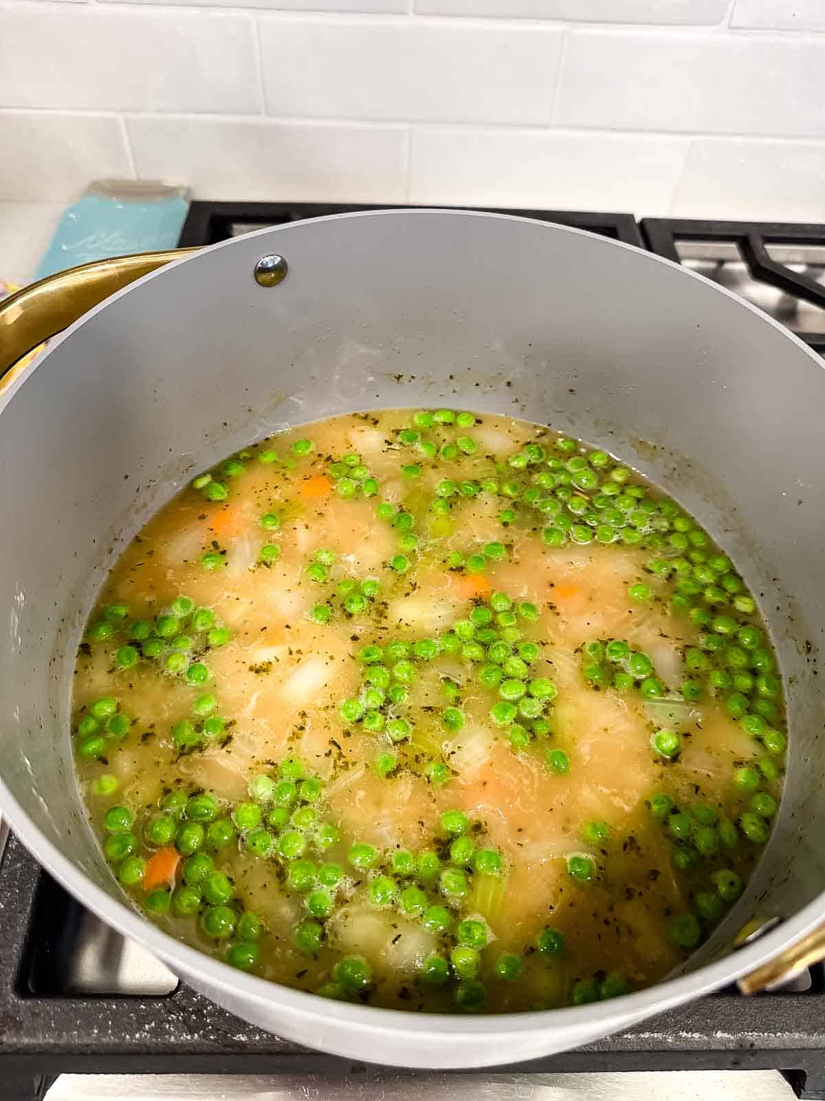 A large pot on a stovetop filled with a soup containing green peas, carrots, potatoes, and herbs in a light broth.
