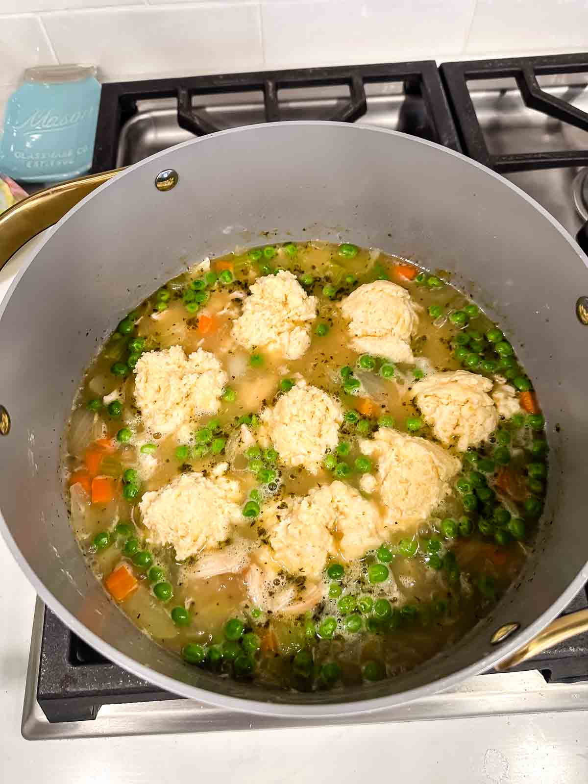 A pot of homemade chicken and vegetable soup simmers on a stove, topped with several raw biscuit dough dumplings, waiting to cook. Visible ingredients include peas, carrots, and broth.