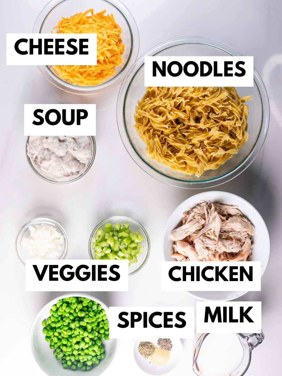 Various ingredients are arranged in bowls on a white surface, each labeled: shredded cheese, egg noodles, creamy soup, chopped veggies, shredded chicken, green peas, spices, and a measuring cup of milk.