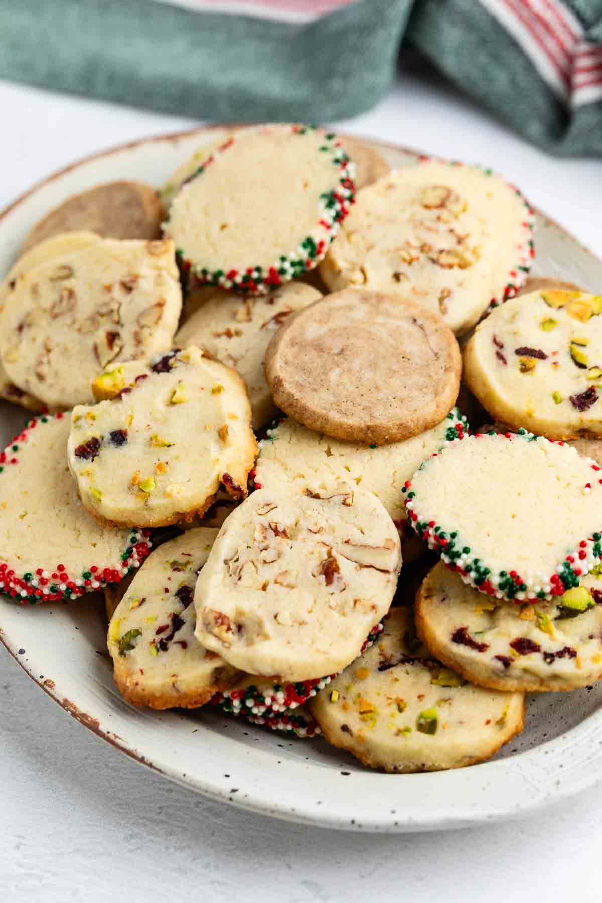 A plate filled with assorted cookies, some with chopped nuts, others with colorful sprinkles on the edges, and a variety of textures and toppings. A cloth napkin is visible in the background.