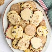 An oval plate filled with assorted slice-and-bake cookies, some with nuts and dried fruit pieces, and others with colored sprinkles on the edges. A green kitchen towel with a red and white stripe is nearby.