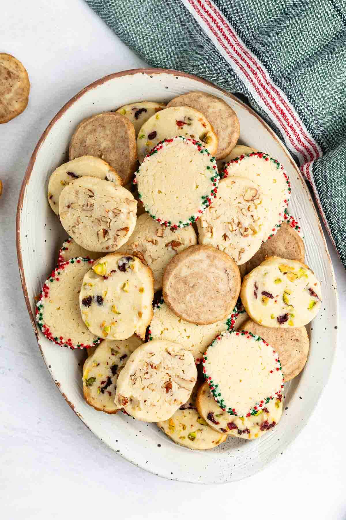 An oval plate filled with assorted slice-and-bake cookies, some with nuts and dried fruit pieces, and others with colored sprinkles on the edges. A green kitchen towel with a red and white stripe is nearby.