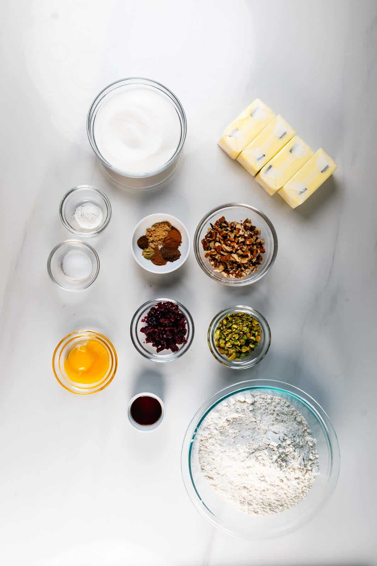 A flat lay of baking ingredients on a white surface, including flour, sugar, butter sticks, an egg, vanilla extract, baking powder, spices, chopped nuts, dried cranberries, and pistachios in small bowls.