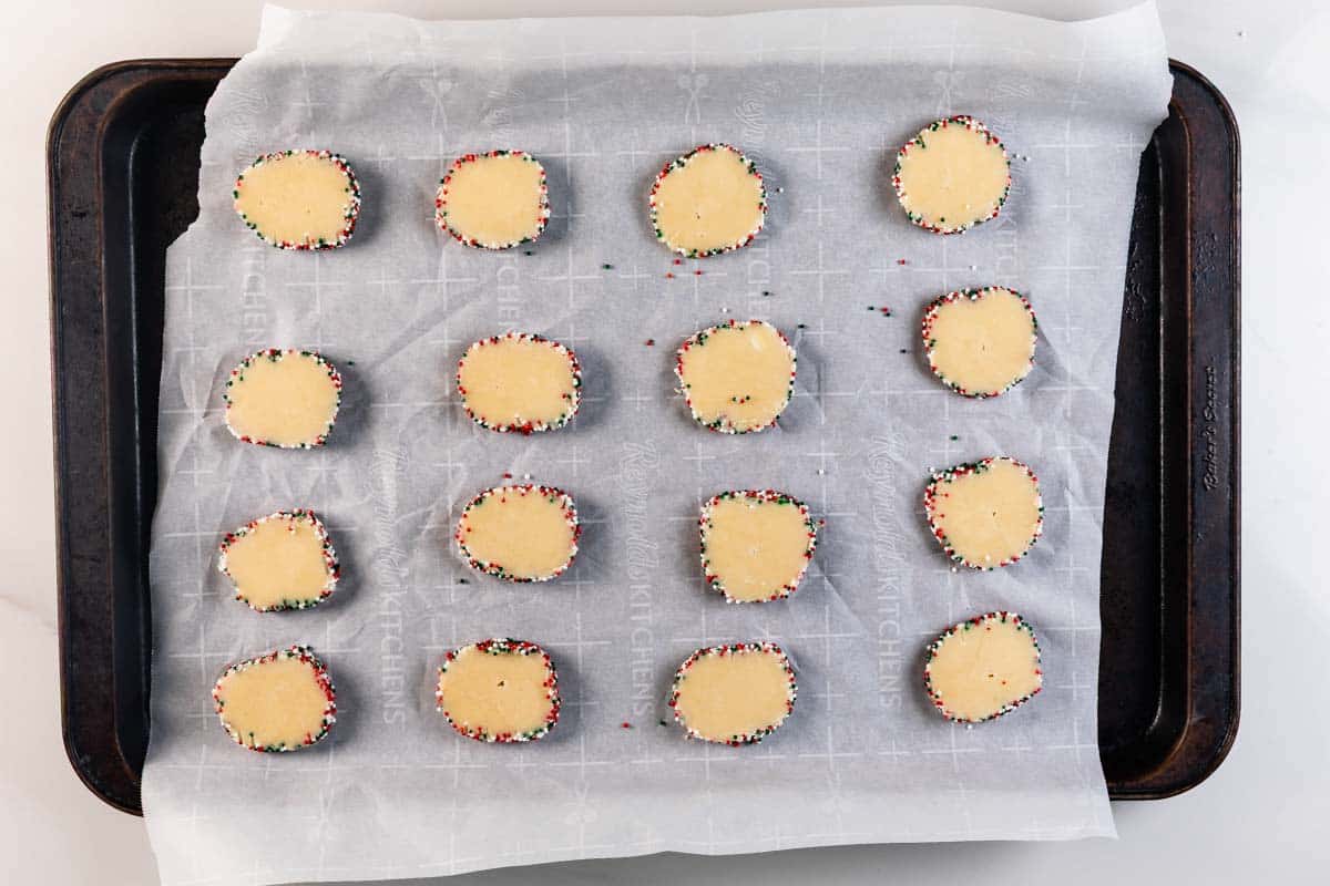 A baking tray lined with parchment paper holds sixteen unbaked square cookies edged with colorful sprinkles, evenly spaced and ready for baking.