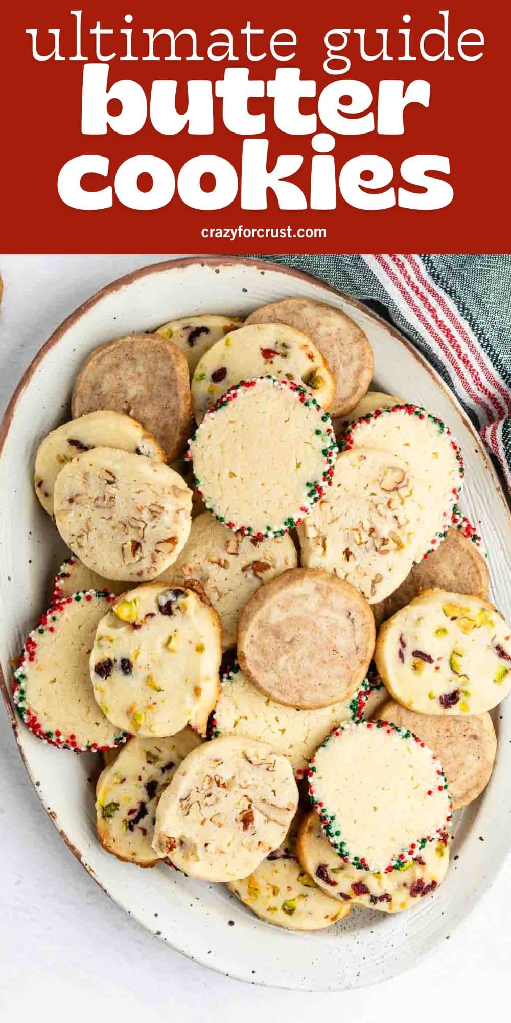 An oval plate filled with various butter cookies, some decorated with colorful sprinkles and others with nuts or dried fruit, sits on a white surface with a red and white striped cloth nearby. Text above reads, ultimate guide butter cookies.