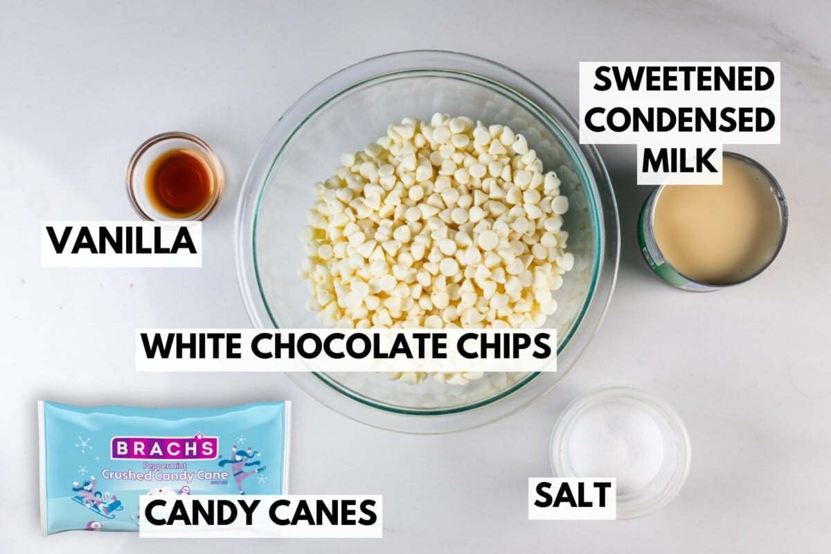 A glass bowl of white chocolate chips surrounded by vanilla extract, sweetened condensed milk, a small bowl of salt, and a bag of Brachs crushed candy canes, all labeled with bold text.