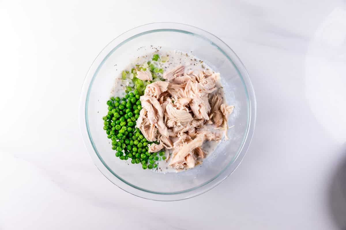 A clear glass bowl on a white surface contains shredded chicken, green peas, and chopped celery mixed with a creamy sauce and black pepper.
