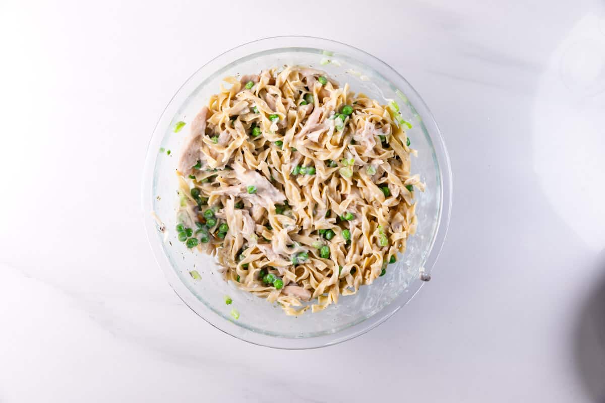 A glass bowl filled with creamy pasta noodles mixed with green peas and chopped green onions, viewed from above on a white surface.