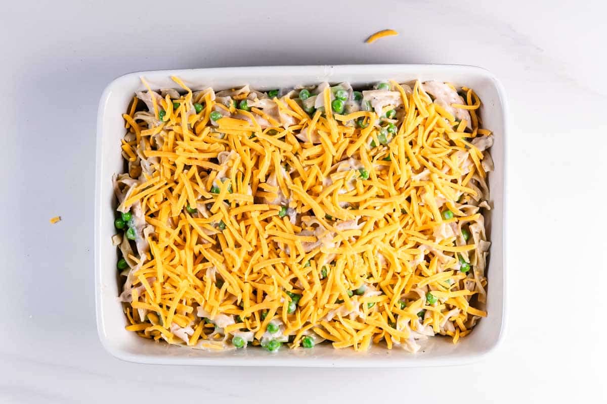 A rectangular baking dish filled with a creamy noodle casserole mixed with peas and topped with a generous layer of shredded cheddar cheese, ready to be baked.