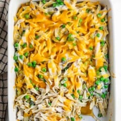 A casserole dish filled with creamy noodle pasta mixed with green peas and topped with melted cheddar cheese, with a serving spoon on the side. A bowl of peas and shredded cheese are nearby.
