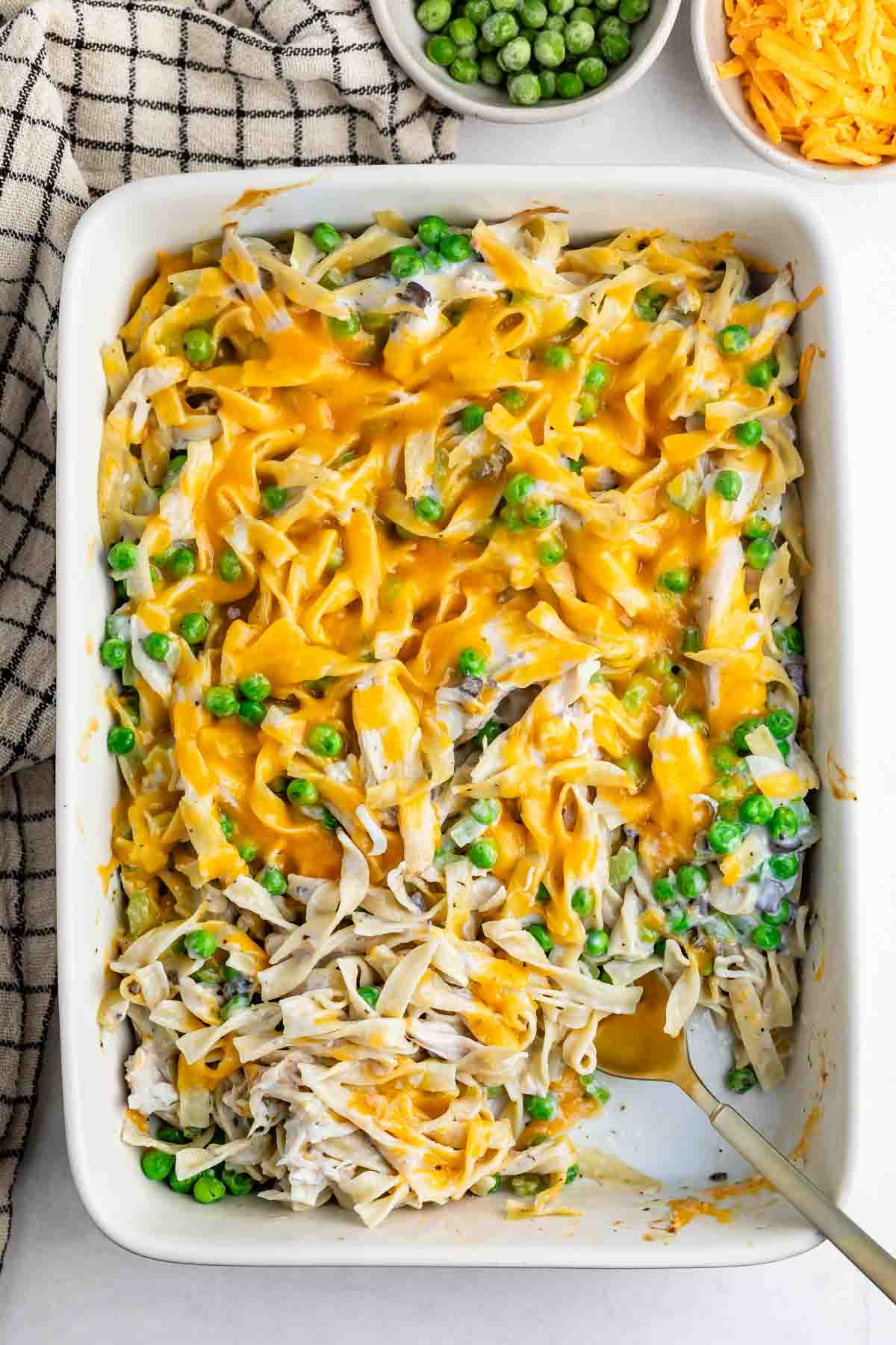 A casserole dish filled with creamy noodle pasta mixed with green peas and topped with melted cheddar cheese, with a serving spoon on the side. A bowl of peas and shredded cheese are nearby.