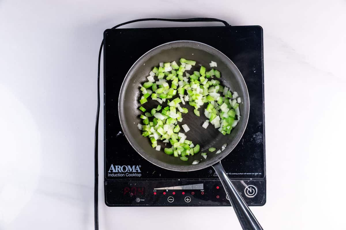 Chopped onions and celery are being saut&eacute;ed in a frying pan on an Aroma induction cooktop, viewed from above.