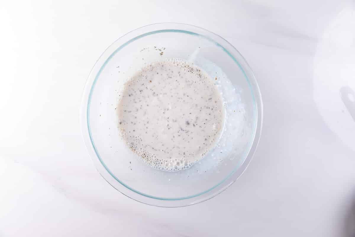 A clear glass bowl filled with a mixture of liquid and small seeds sits on a white marble surface. The mixture appears to be soaking or thickening, possibly chia seeds in milk.