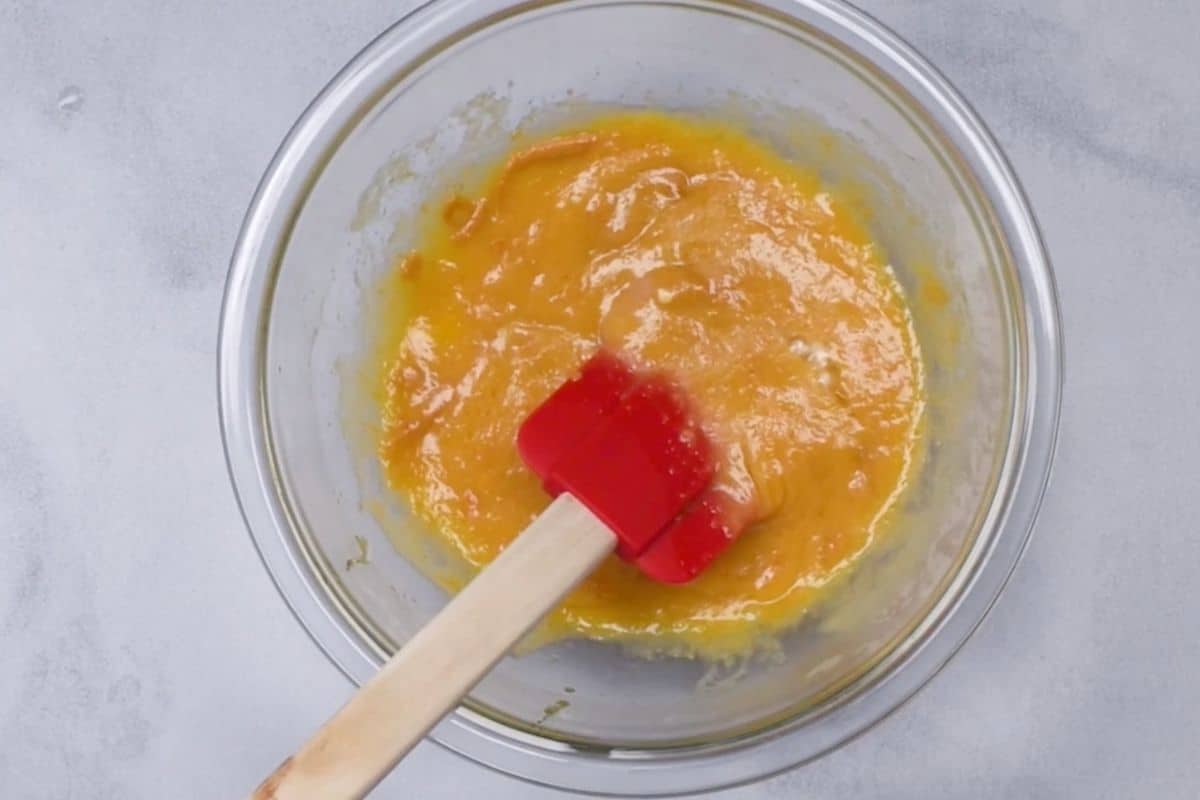 A glass bowl containing a yellow-orange dog cake mixture being stirred with a red spatula on a light gray surface.
