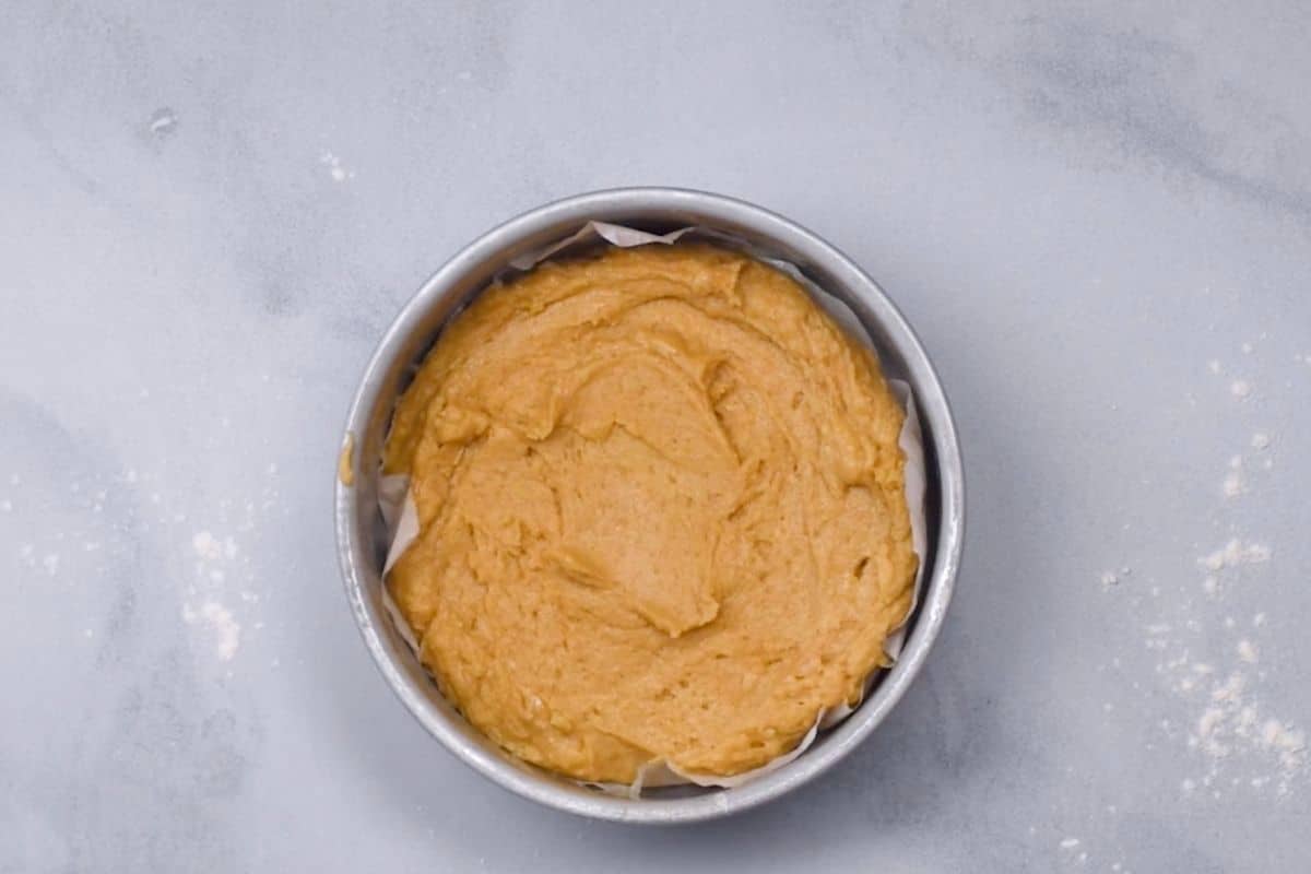 Round metal cake pan filled with unbaked tan dog cake batter on a light gray countertop, with a few scattered flour spots nearby.