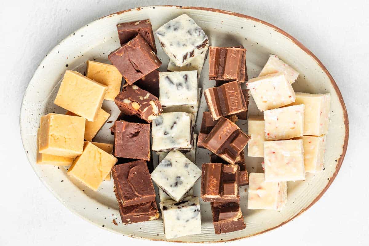 An oval plate with assorted square fudge pieces in different flavors, including light brown, dark brown, white with chocolate bits, and white with pink specks, arranged in groups on a white background.