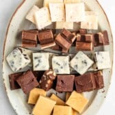 An oval plate holding rows of assorted fudge squares in different flavors and colors, including white, brown, and caramel, neatly arranged on a light background.