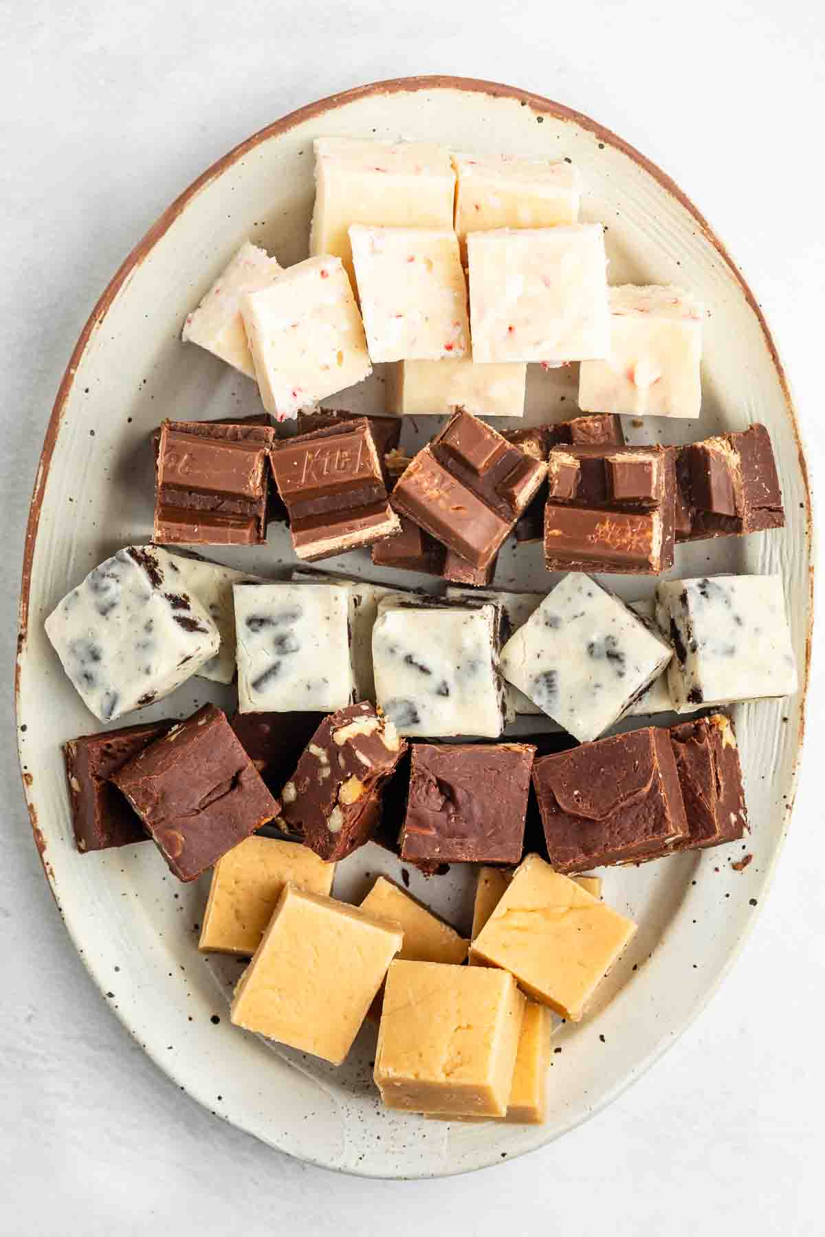An oval plate holding rows of assorted fudge squares in different flavors and colors, including white, brown, and caramel, neatly arranged on a light background.