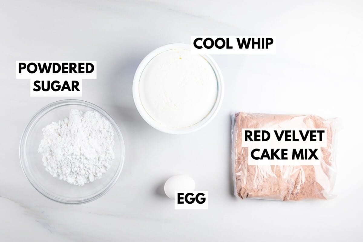 Top-down view of four baking ingredients on a white surface: a bowl of powdered sugar, a container of Cool Whip, a packet of red velvet cake mix, and a single egg. Each item is labeled.