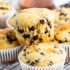 A close-up of chocolate chip muffins in white paper liners, with one muffin broken open to show the inside, all resting on a cooling rack. A glass of liquid is blurred in the background.